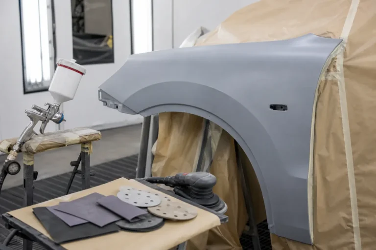 Gray automotive primered fender with spray gun, masking paper, sandpaper sheets, and sanding discs in a paint prep area
